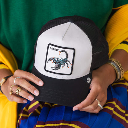 A person holds Goorin Bros.’ black and white mesh “Deadly” hat with a scorpion patch, while wearing colorful clothing plus multiple rings and bracelets.