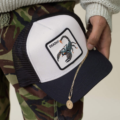 A person holds a Goorin Bros. Deadly white and black mesh hat with a scorpion patch, a gold necklace draped over the hat, and wears camouflage pants.
