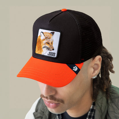 Person wears the Goorin Bros. Good Looks Fox trucker hat—black and orange with a fox patch and "GOOD LOOKS" embroidered—head tilted down in farmyard chic style.