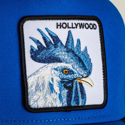 A close-up of the Hollywood Rooster cap by Goorin Bros., featuring a blue design with an embroidered rooster patch and "HOLLYWOOD" above, channeling the iconic red carpet style of the Hills.