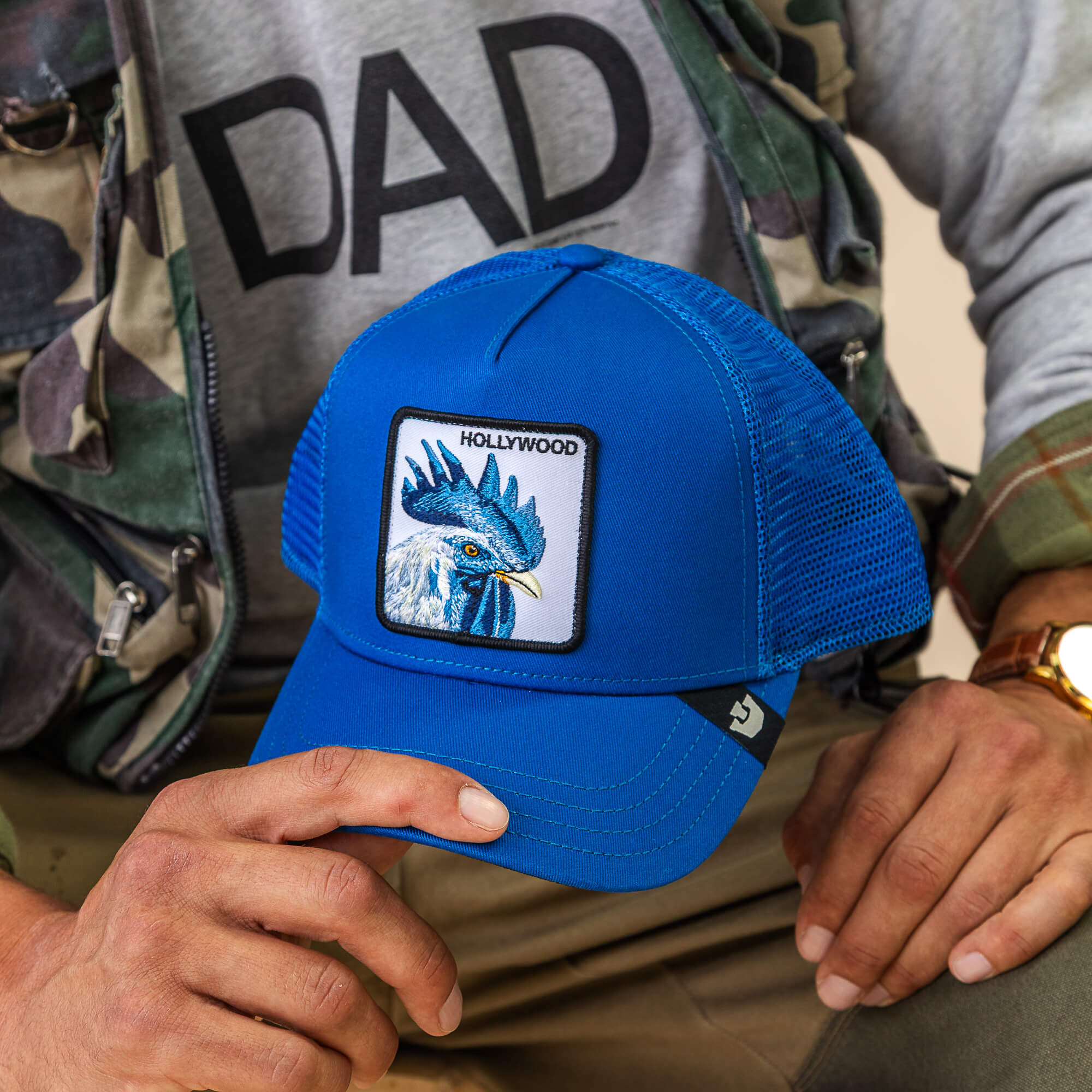 A person holds a blue mesh Goorin Bros. "Hollywood Rooster" cap featuring a blue bird patch and the word HOLLYWOOD, paired with a gray DAD sweatshirt and camo jacket—set for adventure or the red carpet.