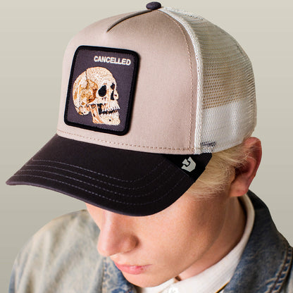 A person wears The Cancelled Skull trucker hat by Goorin Bros., featuring a beige and black design with a stitched skull patch and "CANCELLED" on the front—ideal for bold self-expression.