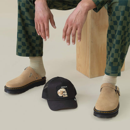 A person in checkered green pants, beige socks, and tan clogs sits on a wooden block. Nearby, the Goorin Bros. "The Cancelled Skull" black trucker hat with skull graphic and "speak your mind" message rests on the floor.