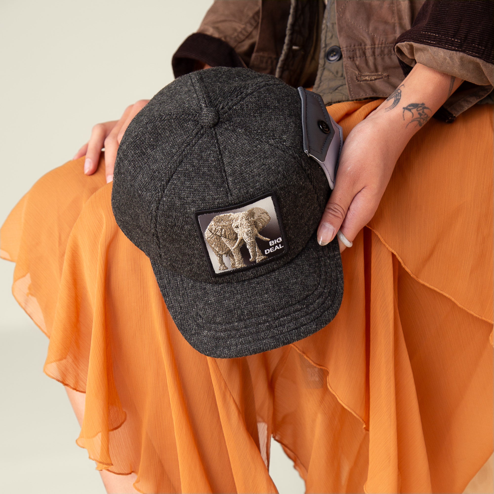A person in an orange skirt holds Goorin Bros.’ Kind of trucker beanie in dark gray, featuring an elephant patch and "BIG DEAL" text, crafted from the ultimate feel-estate for comfort.