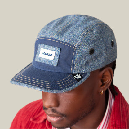 A person wearing a blue denim-textured and navy Billed Demand cap by Goorin Bros., with a visible patch, paired with a red corduroy jacket over a striped shirt, looking slightly downward.
