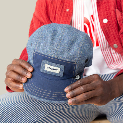 A person in a red jacket and striped pants holds the Billed Demand cap by Goorin Bros., featuring a blue denim texture and a signature Goorin patch on the front.