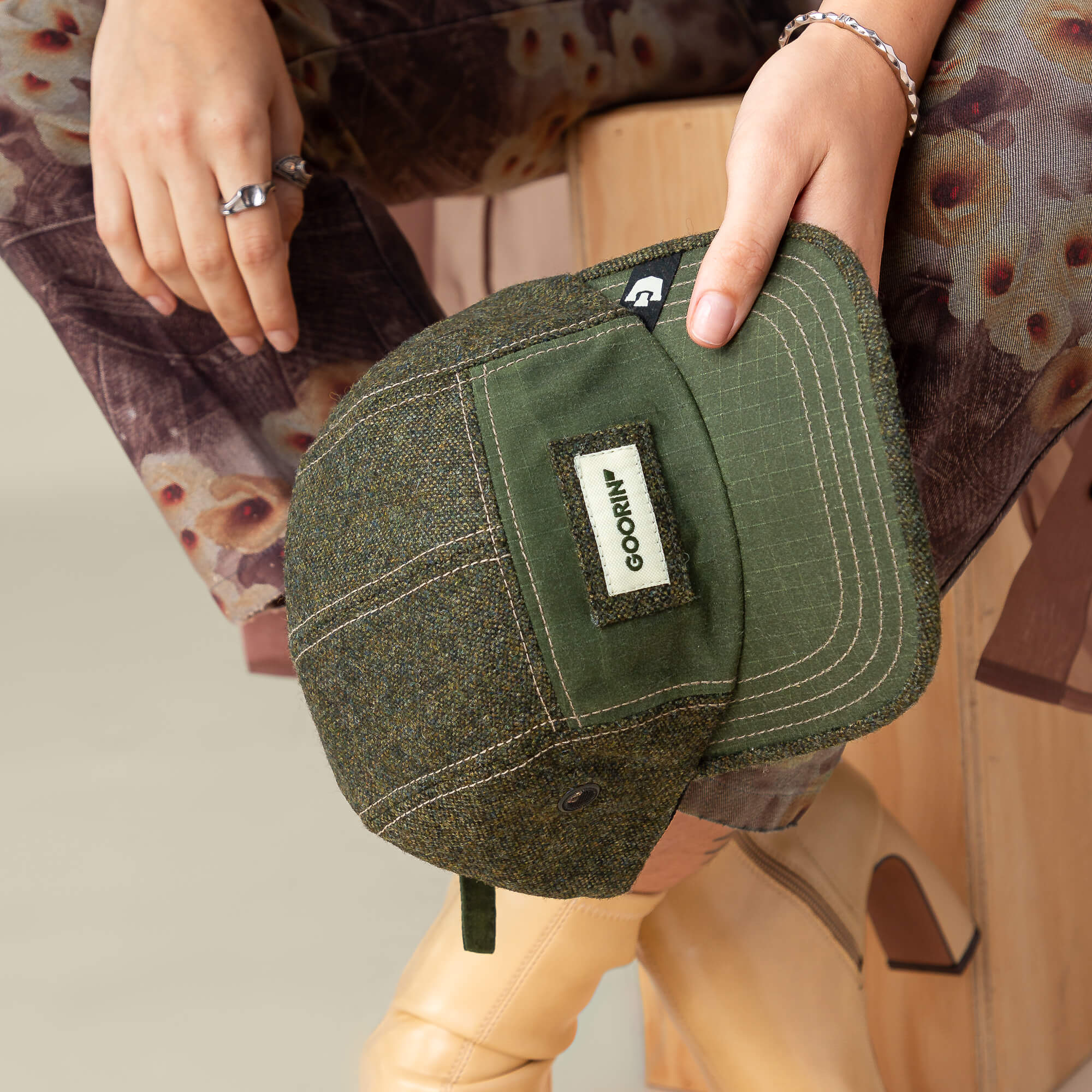 A person sits on a wooden stool holding a green Goorin Bros. Billed Demand cap; they wear floral-patterned pants, beige boots, and silver rings.