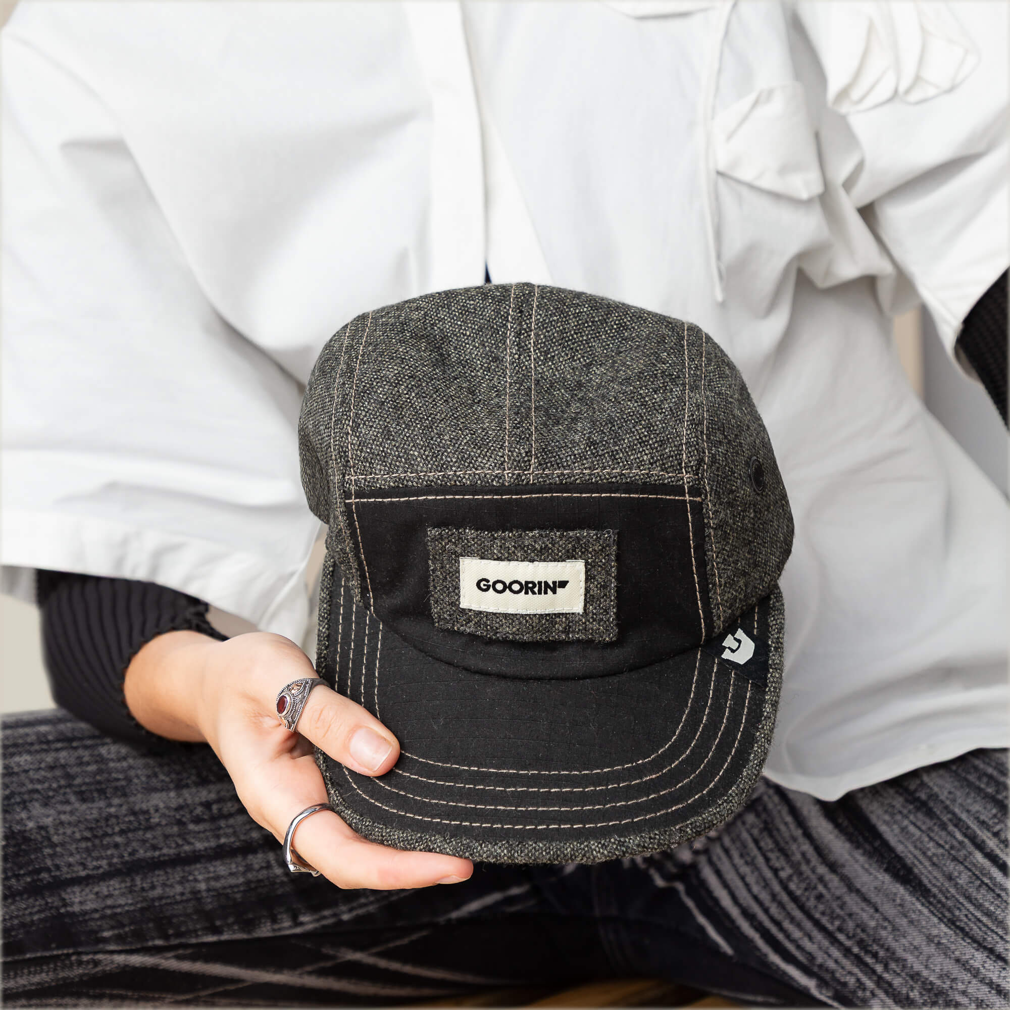 A person in a white shirt and striped pants holds the Billed Demand hat by Goorin Bros., a black denim field hat with a rectangular Goorin patch and distinct denim texture on the front.