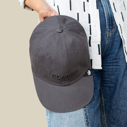 A person holds a Goorin Bros. Papa Core dark gray cap with a soft crown and curved brim, featuring "GOORIN" embroidered on the front, paired with a white patterned shirt and blue jeans.