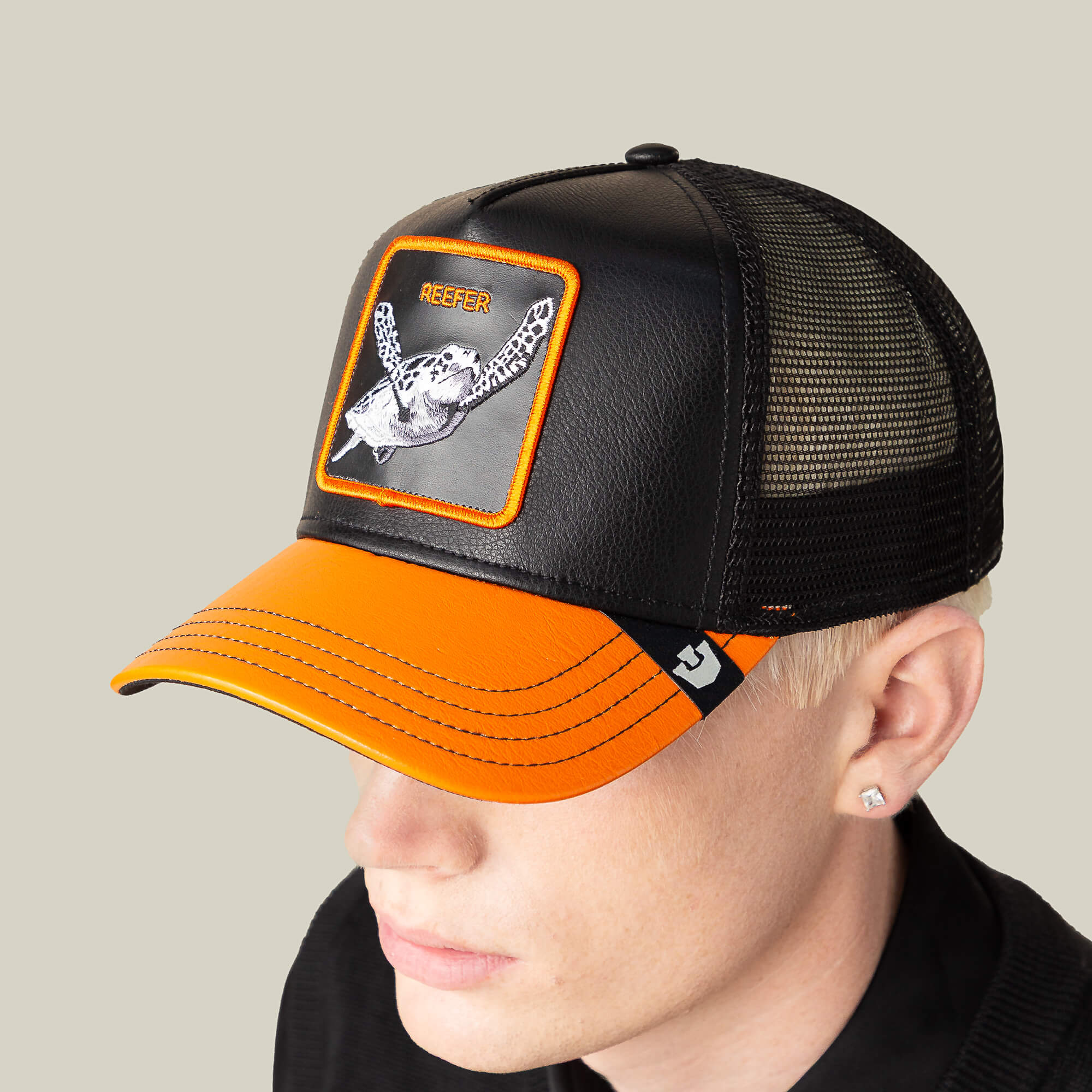 A person with short blonde hair and an earring sports a Goorin Bros. Courtside Reefer trucker hat, featuring a fish patch, black/orange design, and black mesh back, paired with retro high tops for a laid-back look.