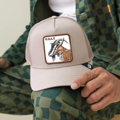 A person holds The G.O.A.T cap by Goorin Bros., a beige hat with a bold goat patch and "G.O.A.T." text, paired stylishly with a green checkered outfit that complements the cap’s neutral tones.