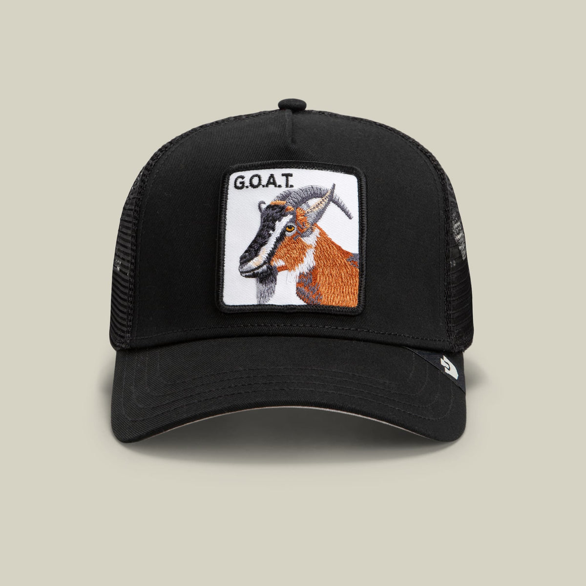 The Goorin Bros. Goat Trucker is a black mesh trucker hat featuring an embroidered white patch with a brown and orange goat design and G.O.A.T. text, displayed against a neutral studio background.