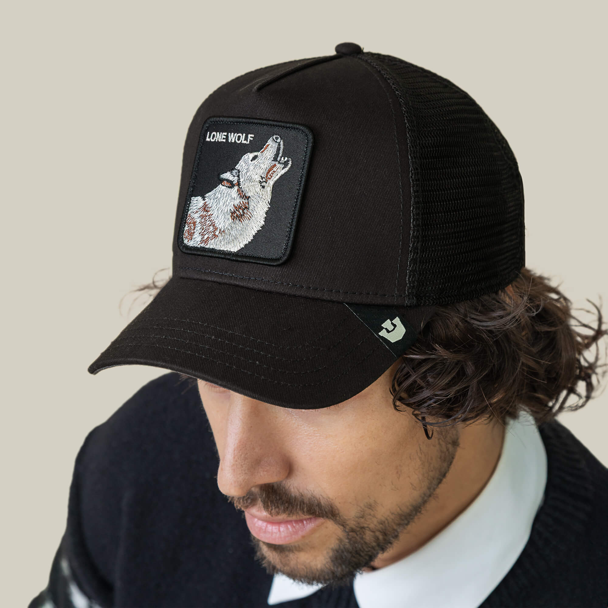 A man wears The Lone Wolf cap by Goorin Bros., featuring a black mesh trucker style and "Lone Wolf" patch with a howling wolf, exuding outlaw energy against a plain beige background.