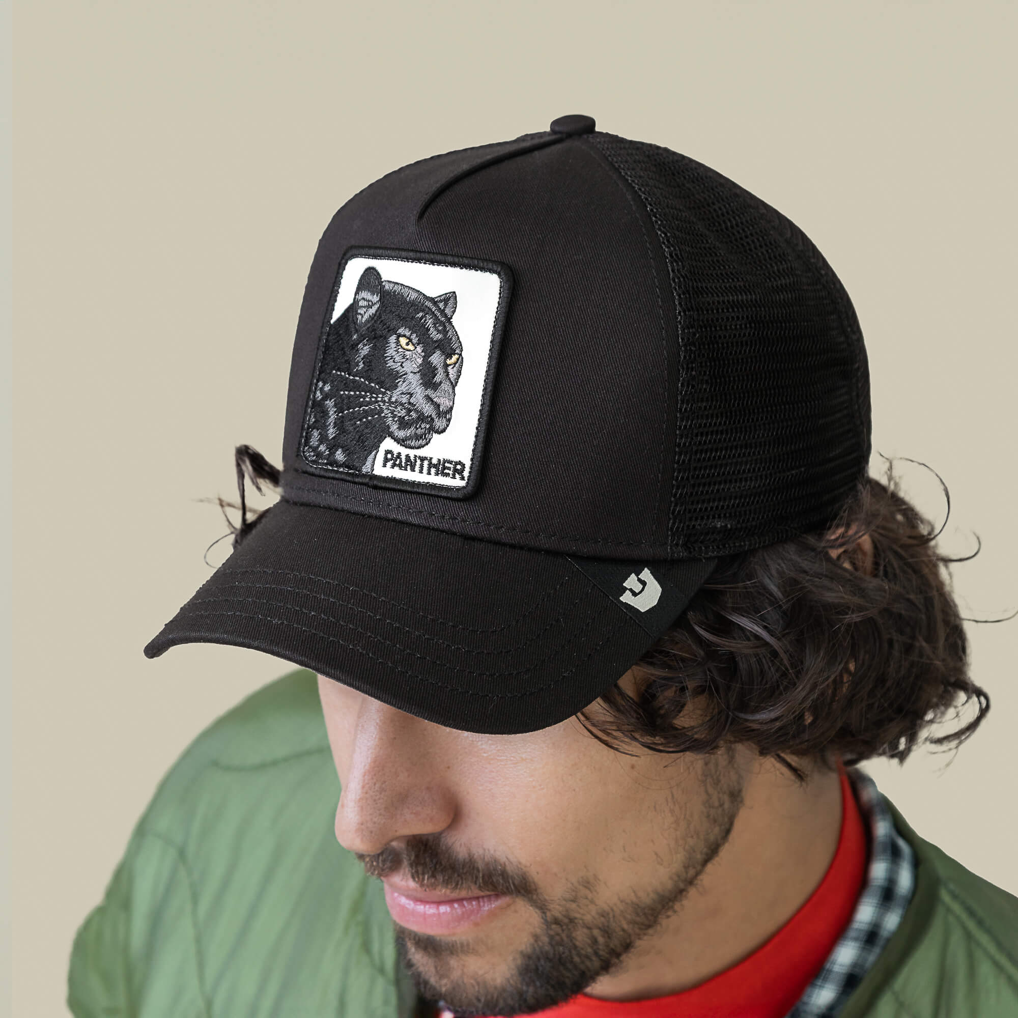 A man in a green jacket and red shirt faces downward, wearing the Goorin Bros. Panther Trucker cap, featuring a black mesh back and panther patch.