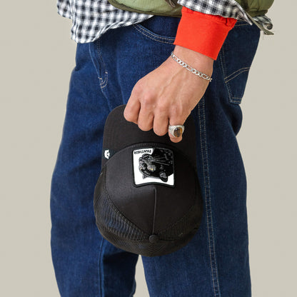 A person holds the Goorin Bros. Panther Trucker cap with a unique patch in their right hand, dressed in blue jeans, a checked shirt with orange sleeves, and silver jewelry.