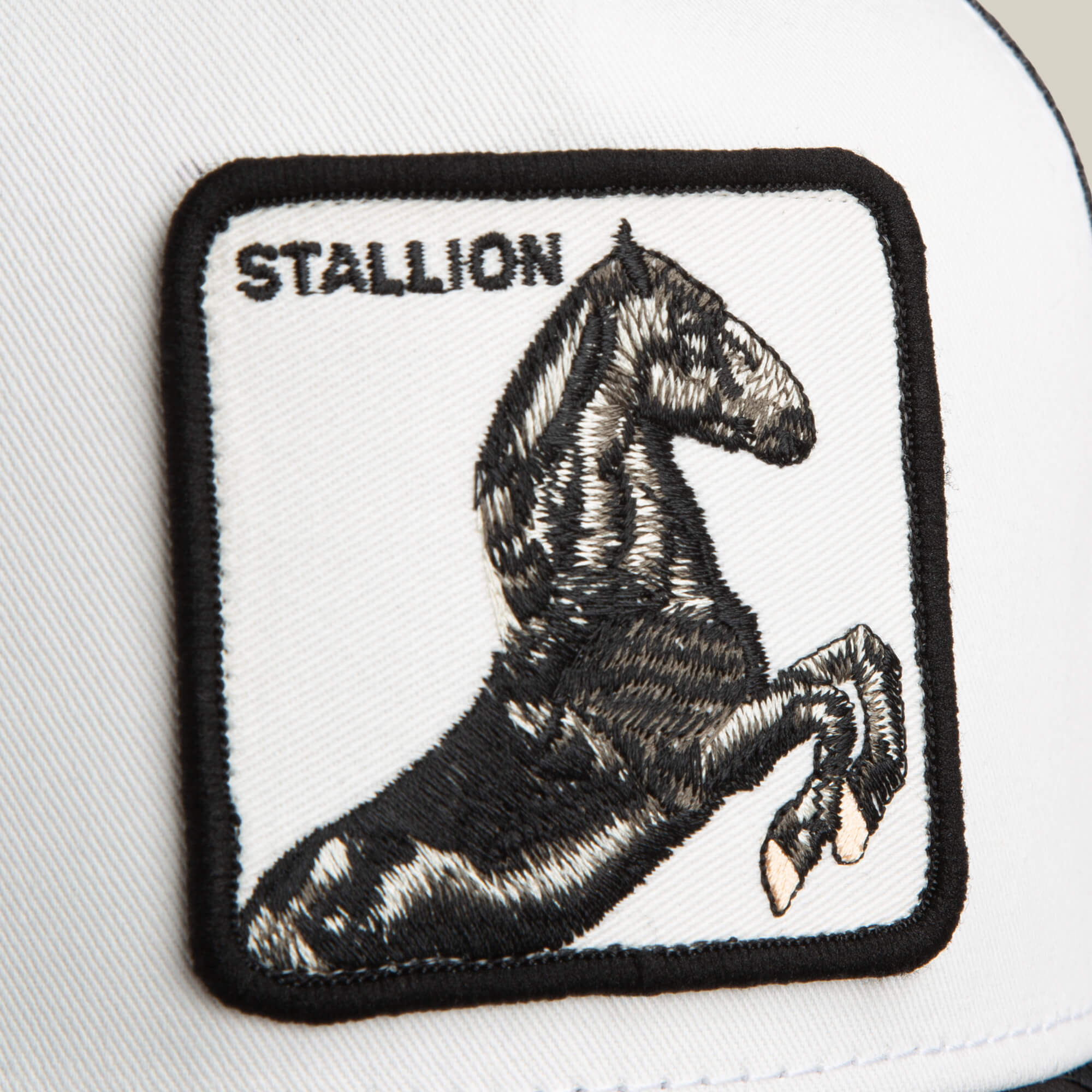 Close-up of the Goorin Bros. Stallion Trucker, a black and white hat showcasing a square patch with an embroidered black stallion and the word "STALLION" at the top left.