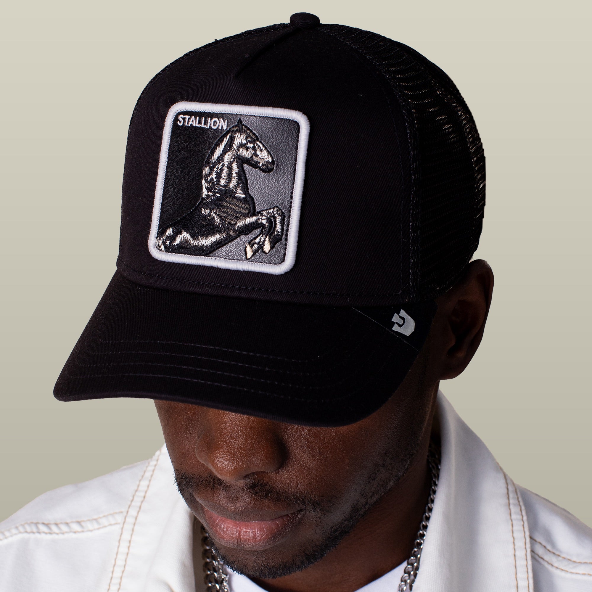 A person wearing the Goorin Bros. Stallion Trucker hat—black and white with a horse patch—paired with a white jacket and a silver chain necklace.