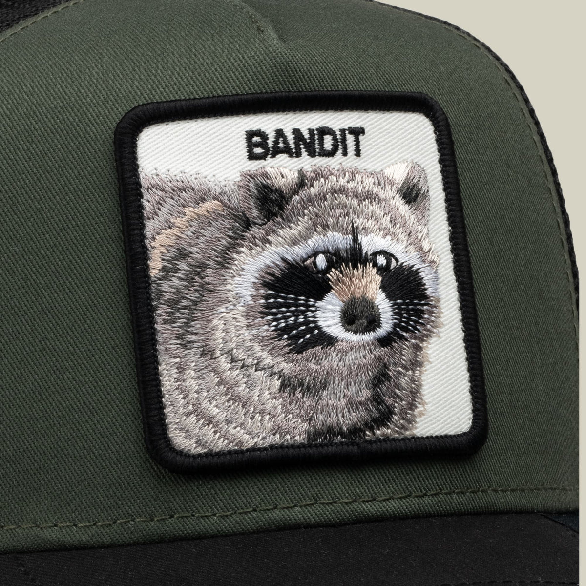 The Goorin Bros. Bandit Trucker is a sage green and black trucker hat featuring an embroidered raccoon patch with "BANDIT" text on the front panel, displayed in close-up against a neutral background.