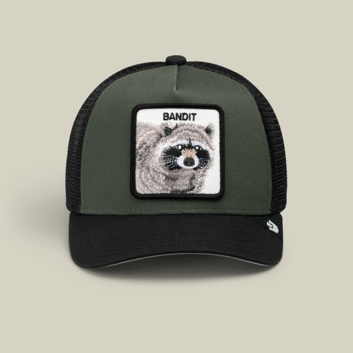 The Goorin Bros. Bandit Trucker is an olive green and black colorblock trucker hat with a mesh back and snapback closure, featuring a rectangular patch with a detailed raccoon illustration and the word Bandit on the front panel.