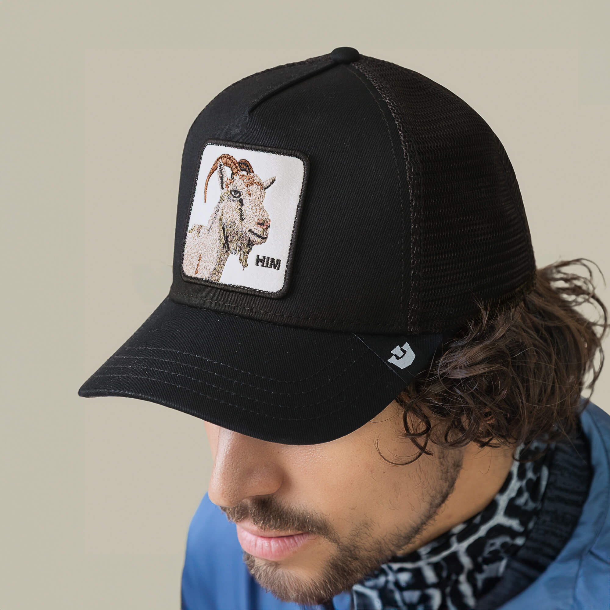 A person wears the Mirage HIM black mesh trucker hat by Goorin Bros., featuring a GOAT illustration patch on the front panel and a stitched warning label for added flair.