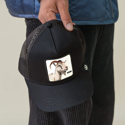 A person holds a black mesh Mirage HIM cap from Goorin Bros., featuring a stitched warning label and a patch with an embroidered GOAT and the word "HIM.