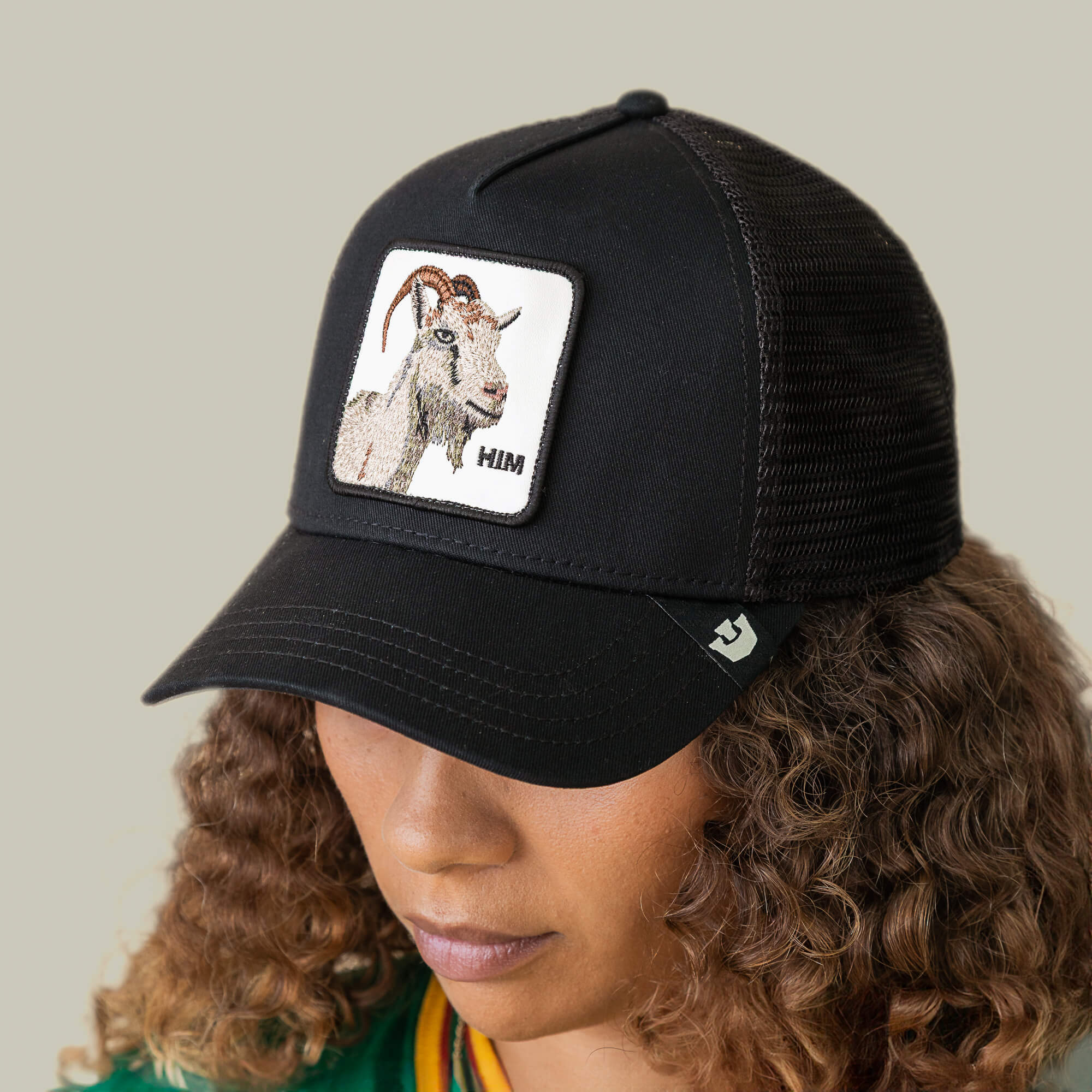 A person wearing a black mesh Mirage HIM trucker hat by Goorin Bros., featuring a patch with an illustration of a GOAT and the word HIM.