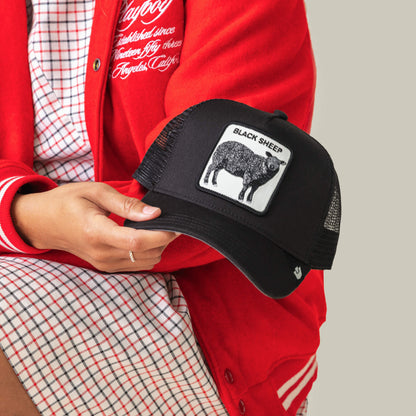 A person in a red jacket and plaid dress holds The Black Sheep cap by Goorin Bros., a black mesh hat featuring a patch with a black sheep and the text "BLACK SHEEP"—ideal for standing out.