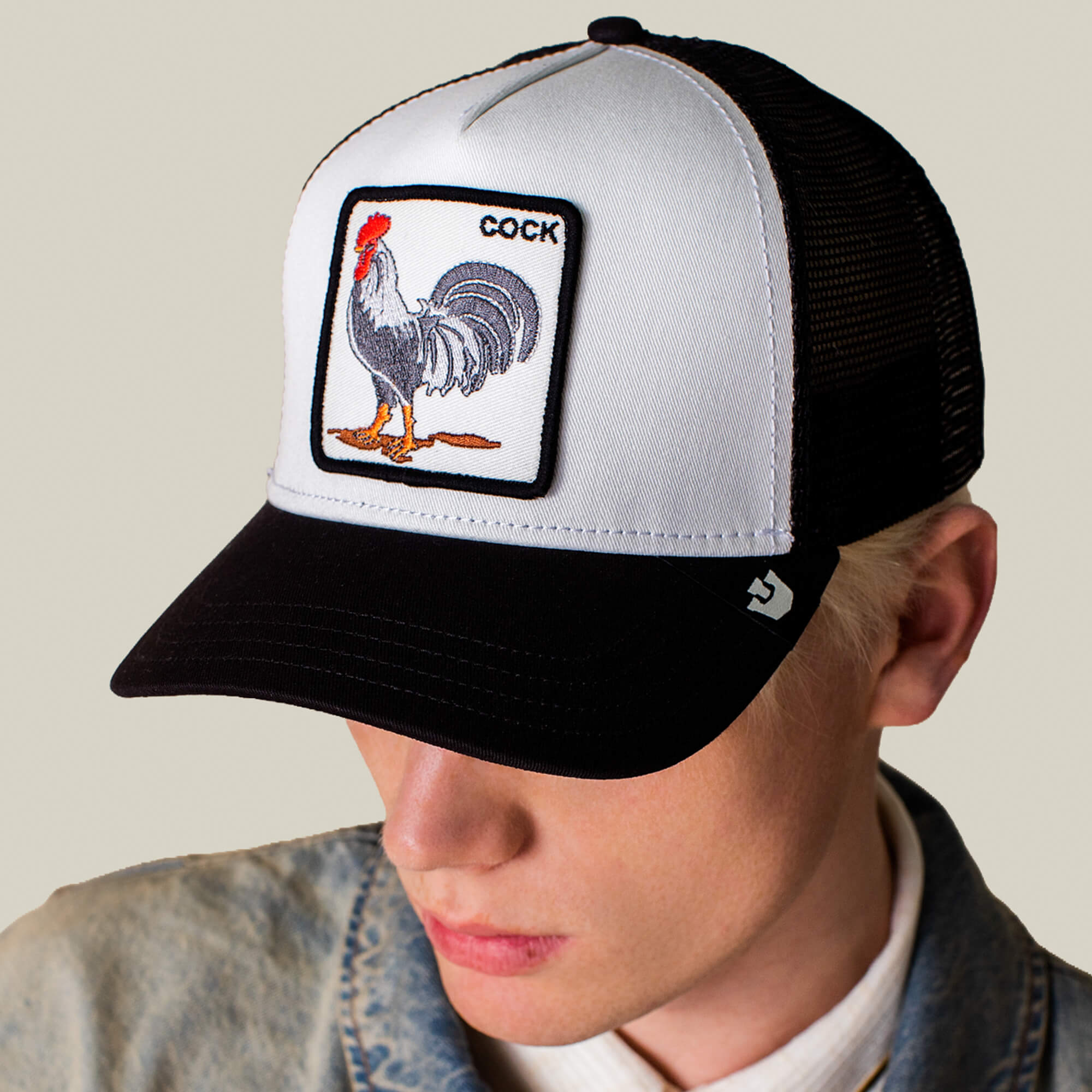 A person wearing The Rooster trucker hat by Goorin Bros., featuring a black and white design with a patch of a rooster and the word “COCK.”.