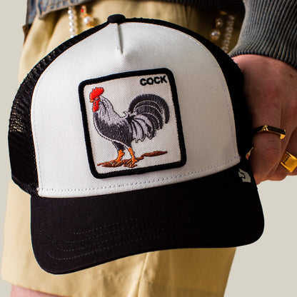 A person holds "The Rooster" hat by Goorin Bros., a black and white trucker cap with a bold rooster patch and the word "COCK" above it.