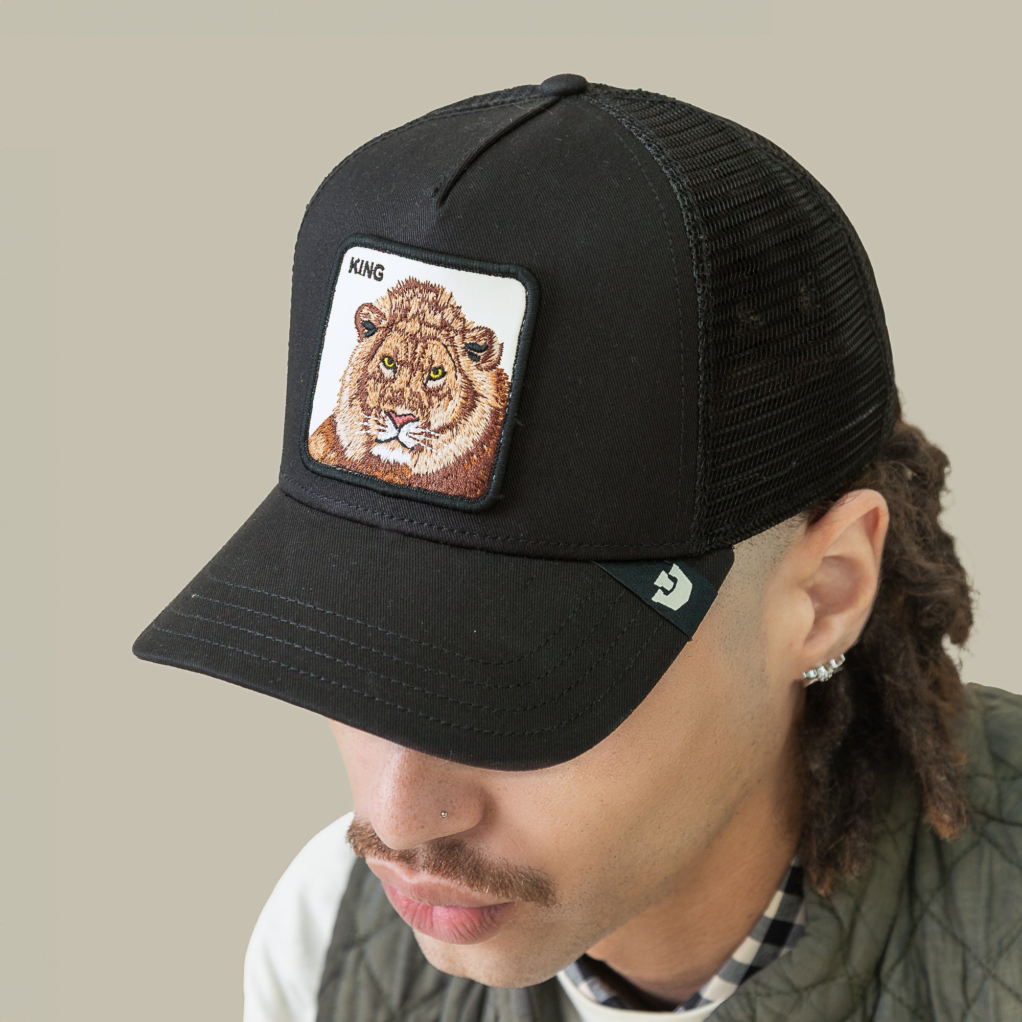A person in the Goorin Bros. King Trucker cap, featuring a bold lion patch and "KING" on the front, sports facial hair and looks down, embracing power dressing with this striking black mesh accessory.