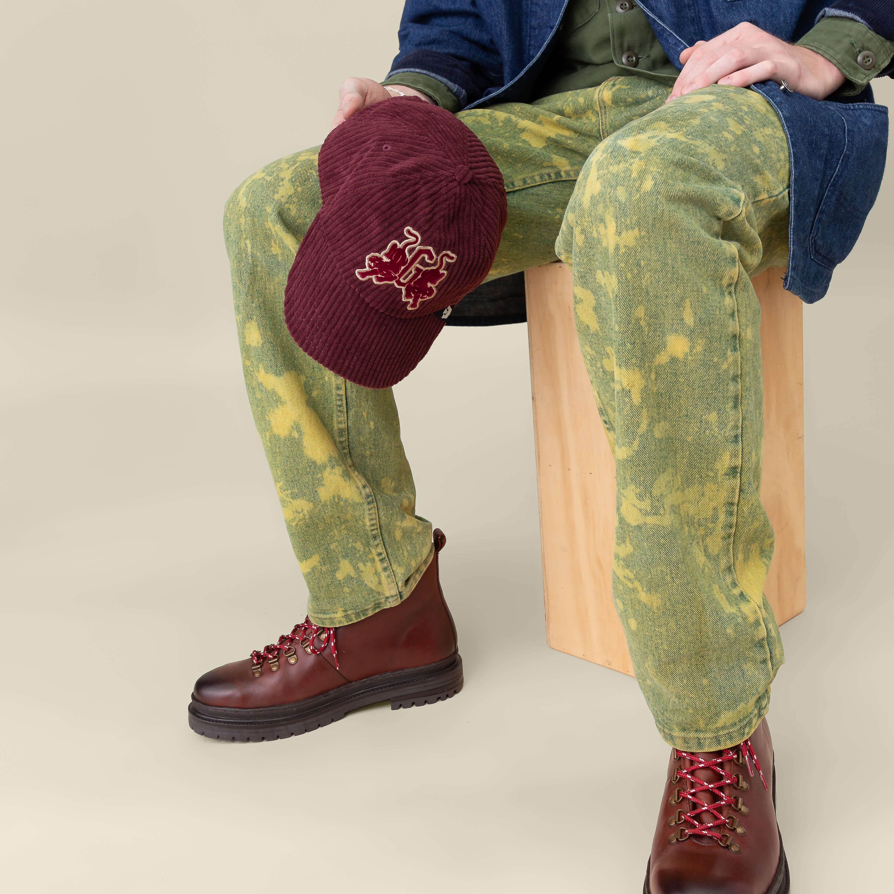 A person seated on a wooden stool wearing yellow-green patterned jeans, brown boots with red laces, and holding the Goorin Bros. Bleacher Report cap with an embroidered corduroy crown.