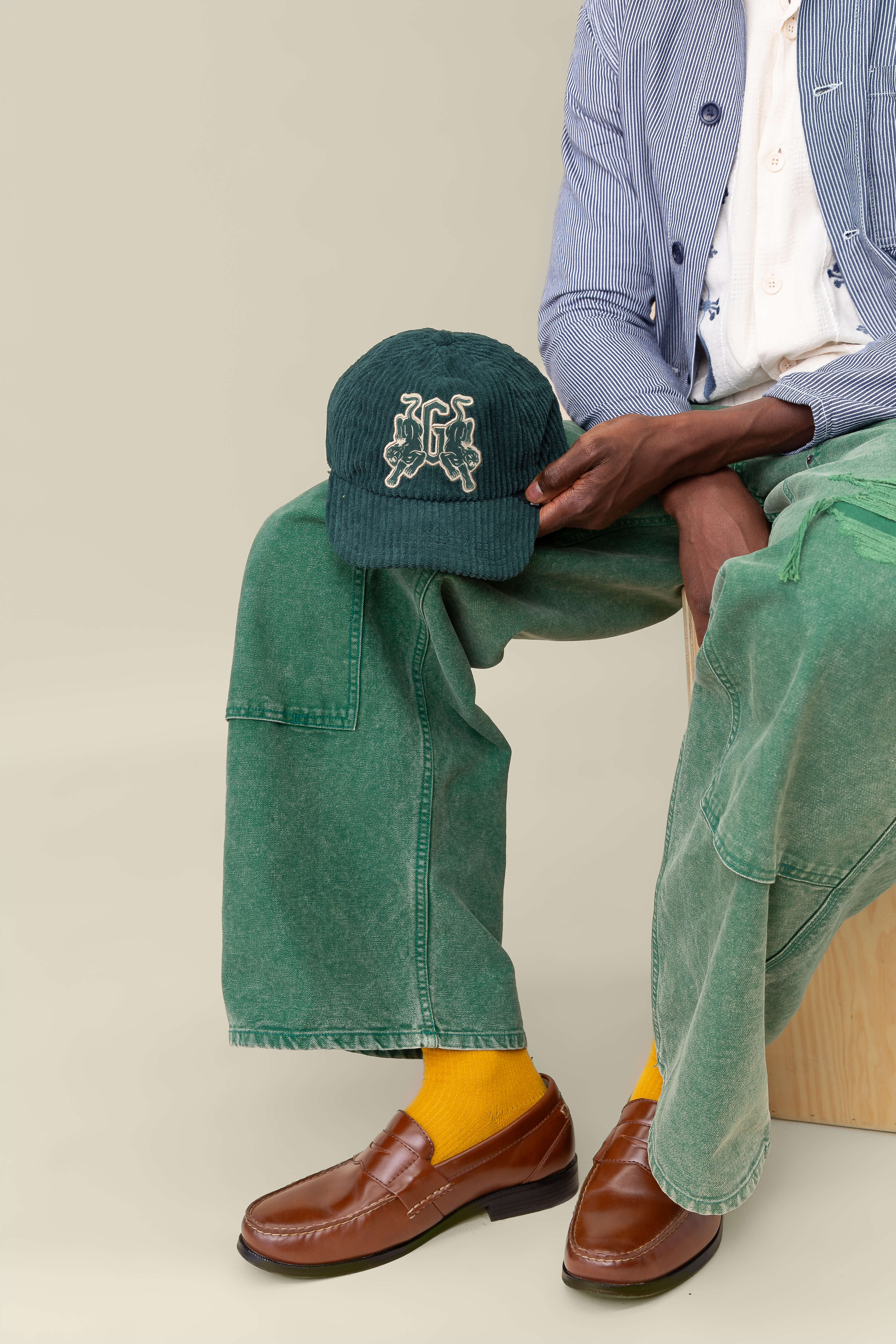 A person sits on a wooden stool in green pants, a blue-striped jacket, brown loafers, yellow socks, and holds the Goorin Bros. Bleacher Report cap—a green corduroy Papa Cap featuring an embroidered logo.