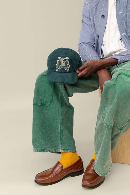 A person sits on a wooden stool in green pants, a blue-striped jacket, brown loafers, yellow socks, and holds the Goorin Bros. Bleacher Report cap—a green corduroy Papa Cap featuring an embroidered logo.