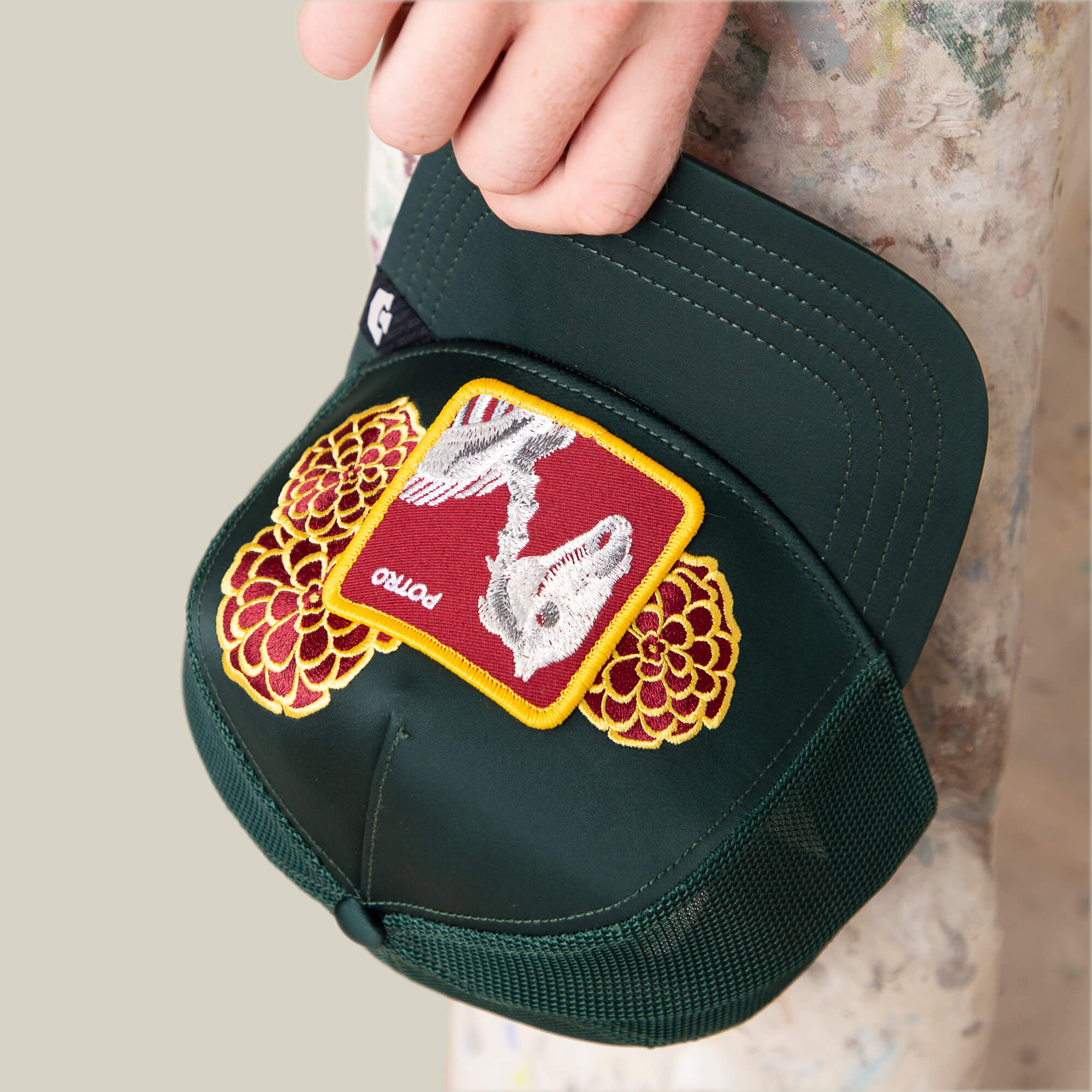A hand holds the Goorin Bros. Dia De Potro cap—dark green with mesh sides, bright yellow and red floral embroidery, and a patch featuring a skeleton on a red background labeled "potto.