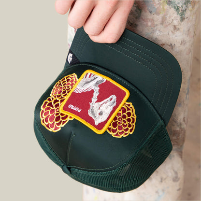 A hand holds the Goorin Bros. Dia De Potro cap—dark green with mesh sides, bright yellow and red floral embroidery, and a patch featuring a skeleton on a red background labeled "potto.