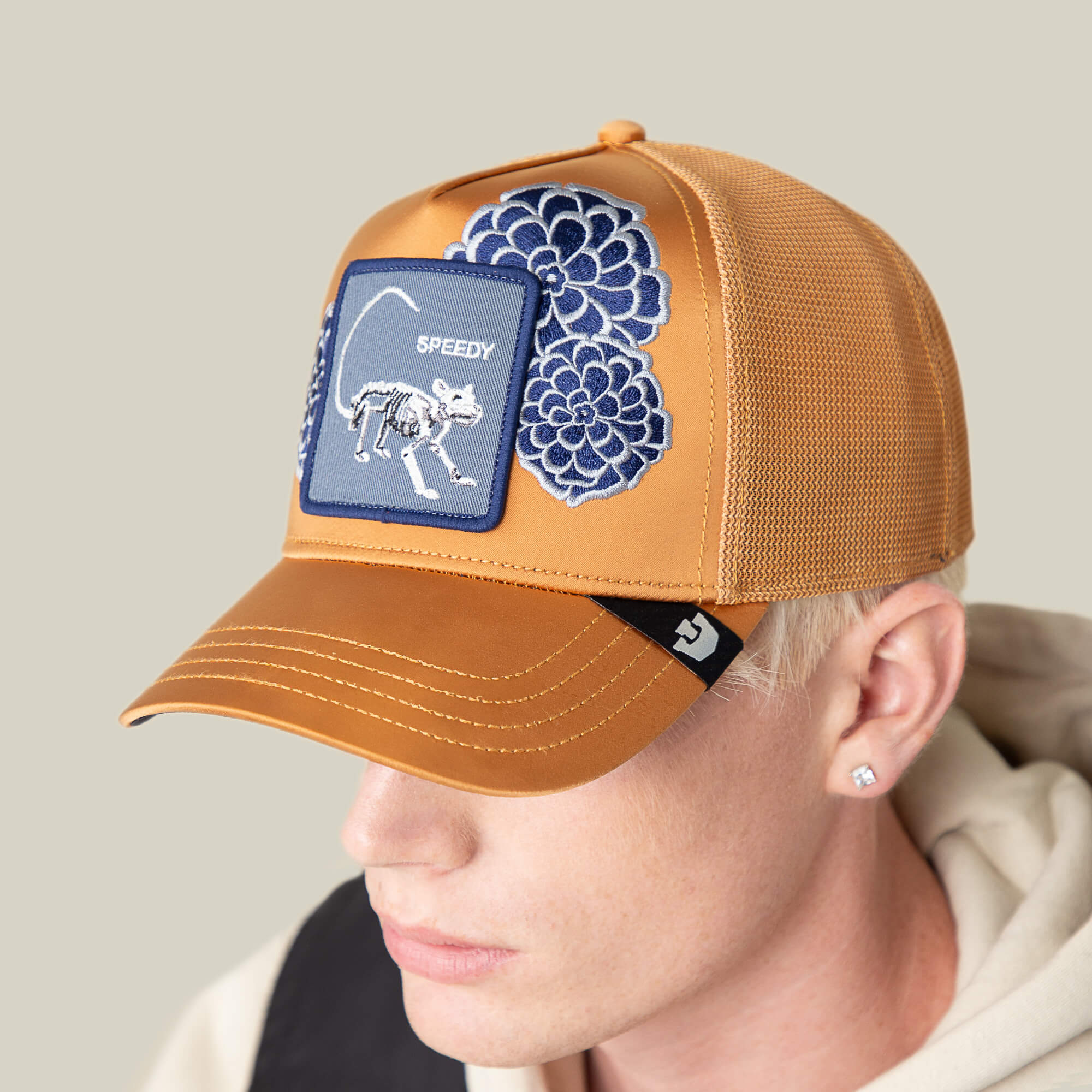 A person wearing the Goorin Bros. Dia De Speedy tan mesh baseball cap, featuring blue floral embroidery on the crown and a patch with a cat skeleton and "SPEEDY.