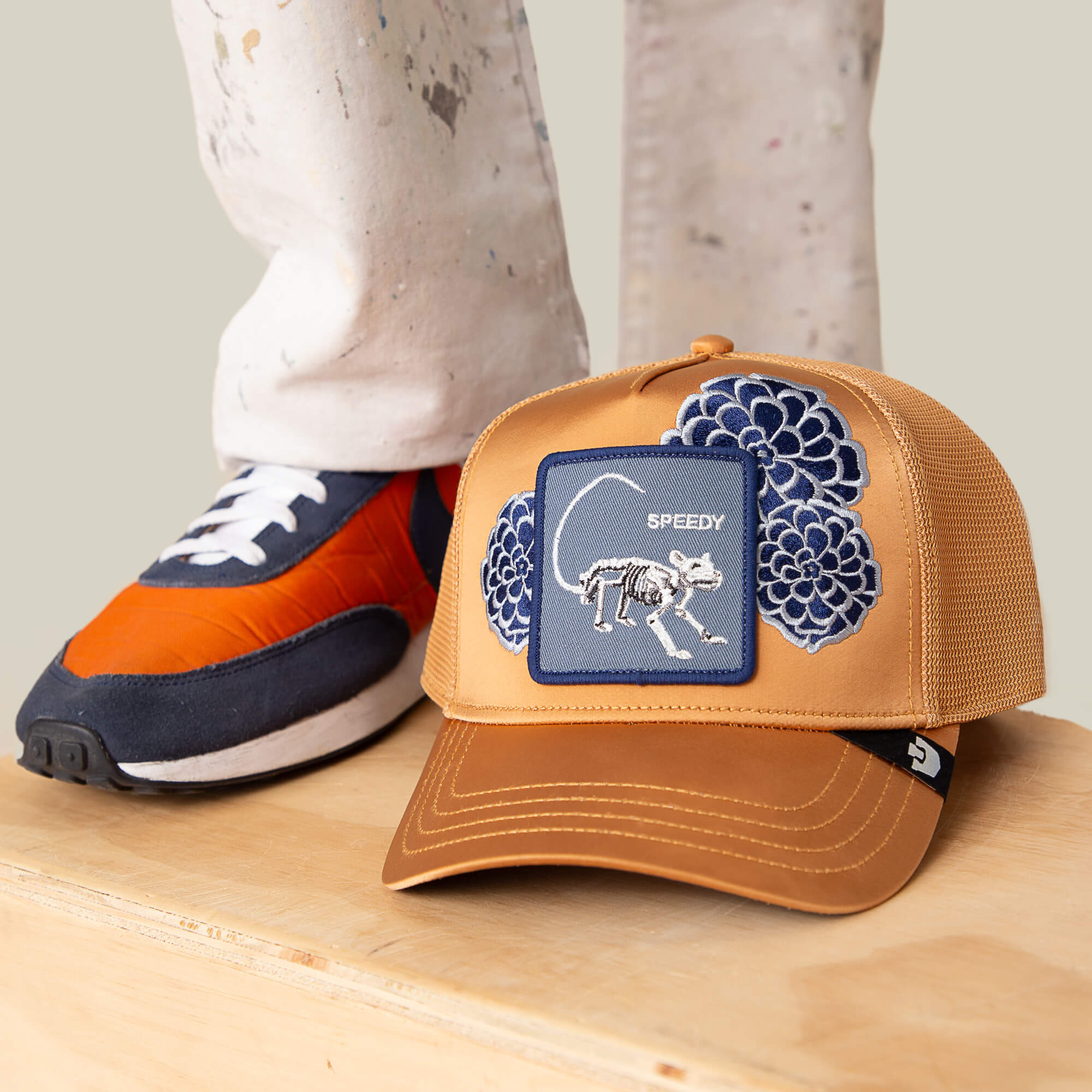 The Dia De Speedy trucker hat by Goorin Bros. features tan fabric, blue "fearless" embroidery, and a SPEEDY skeleton patch, displayed on a wooden surface beside someone in white pants and colorful sneakers.