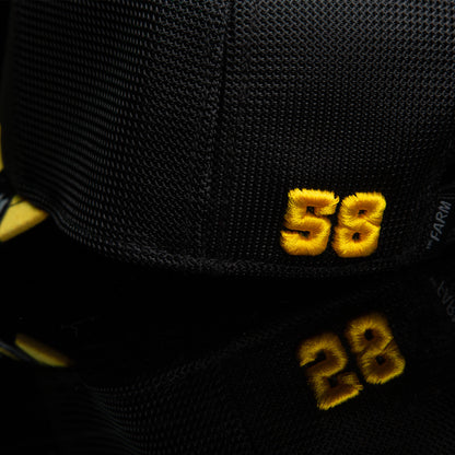 Close-up of the Goorin Bros. Suede Tanger 58 black mesh cap with yellow embroidery, reflected on a glossy black surface.