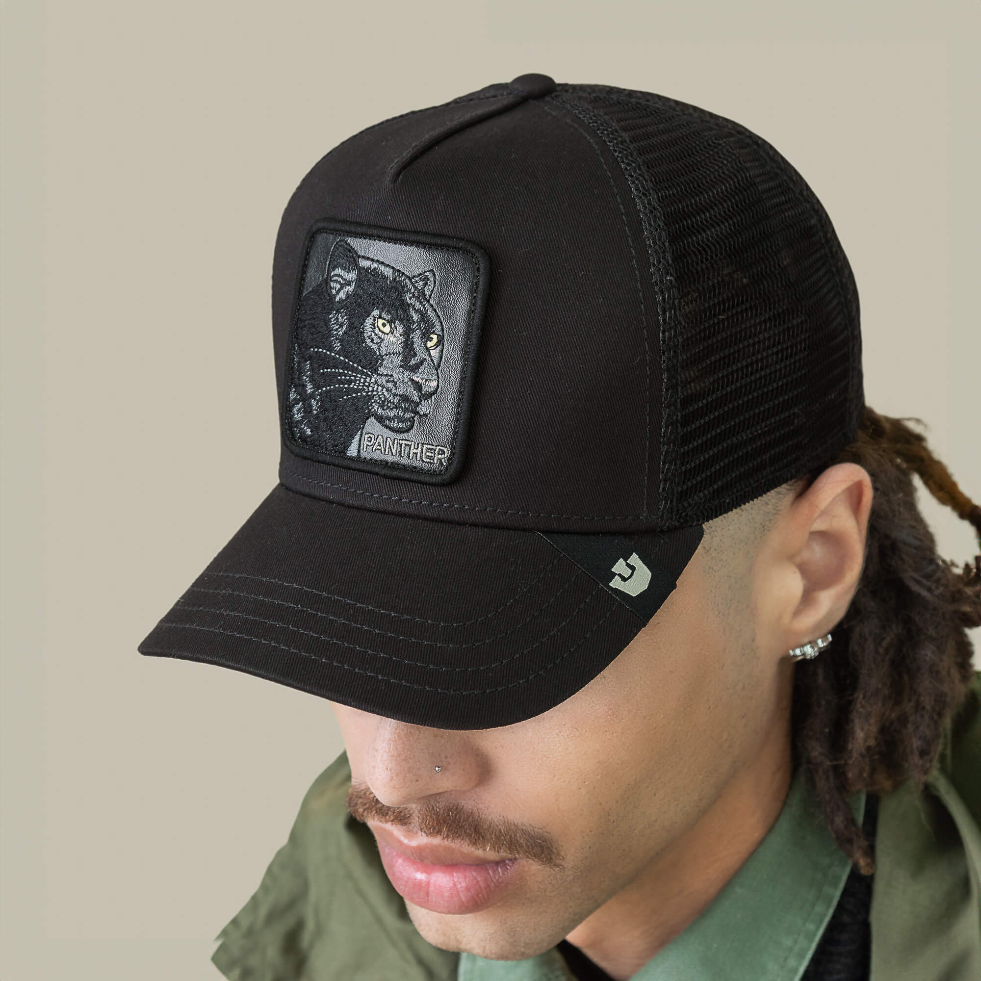 A person wearing The Panther by Goorin Bros.—a black mesh baseball cap featuring a panther patch—paired with a green jacket and green collared shirt.