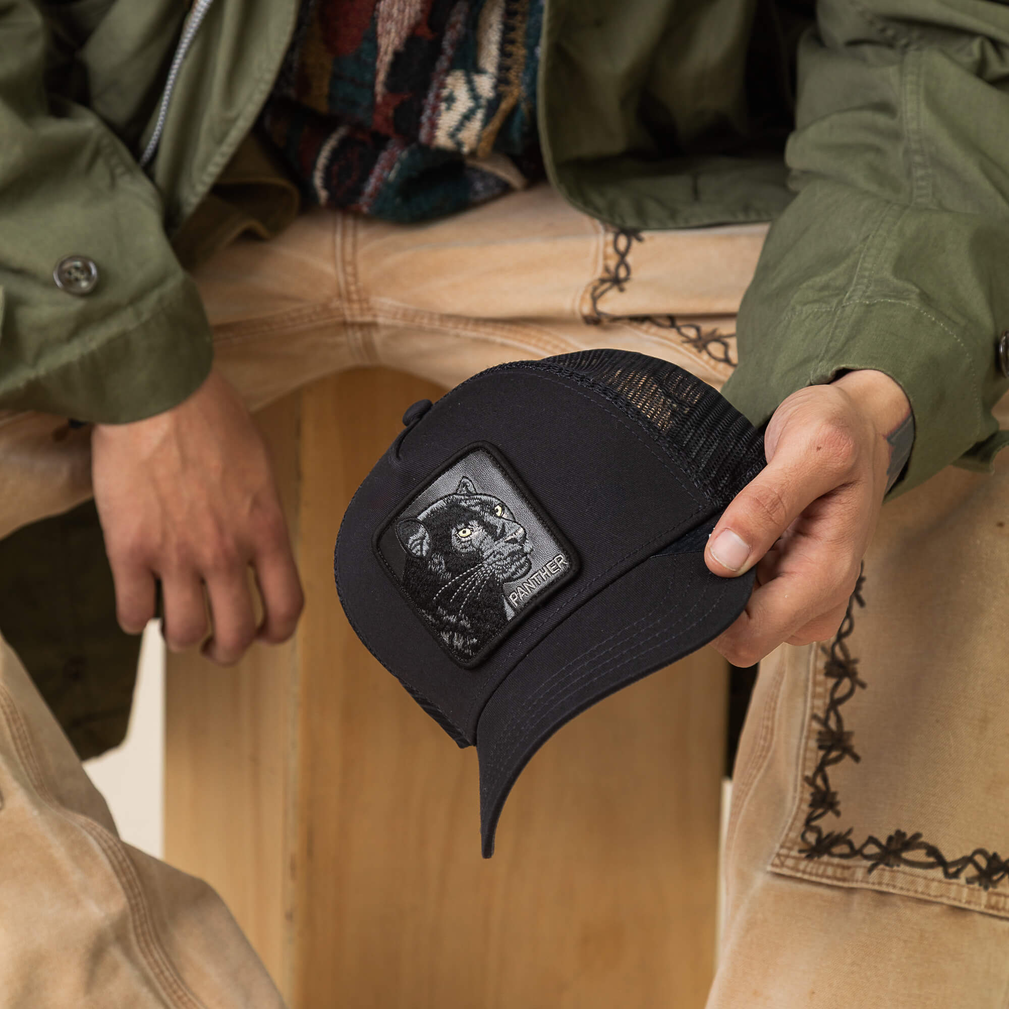 A person sits on a wooden crate, holding Goorin Bros.' The Panther hat featuring a black cat patch, and wearing tan embroidered pants with a green jacket.