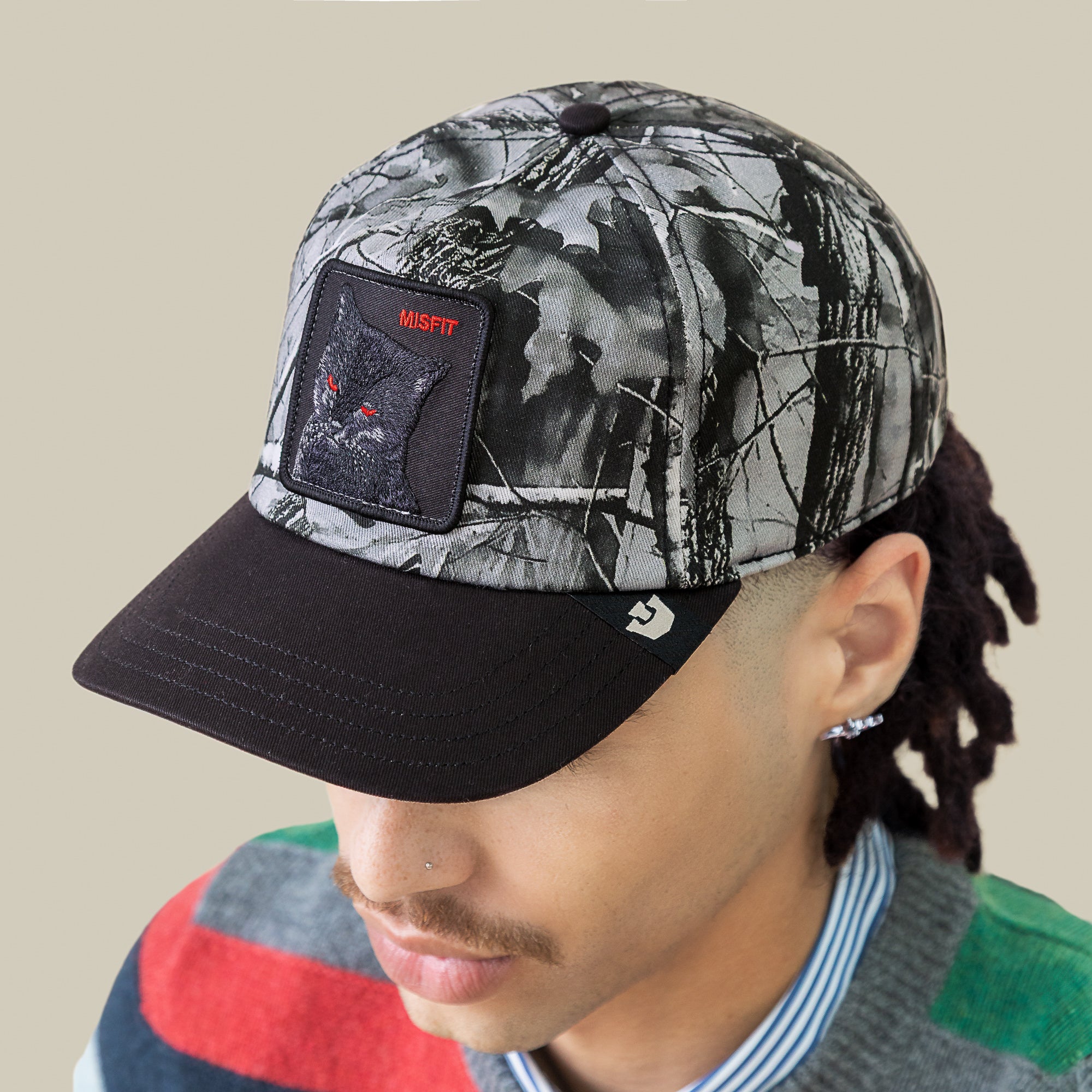 A person wears the Goorin Bros. Misfit Camo cap, featuring a wolf patch labeled "MISFIT." The Low Pro Fit style is paired with a mustache and a vibrant sweater layered over a striped shirt.