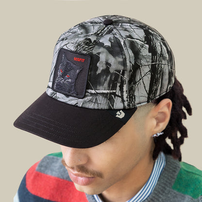 A person wears the Goorin Bros. Misfit Camo cap, featuring a wolf patch labeled "MISFIT." The Low Pro Fit style is paired with a mustache and a vibrant sweater layered over a striped shirt.