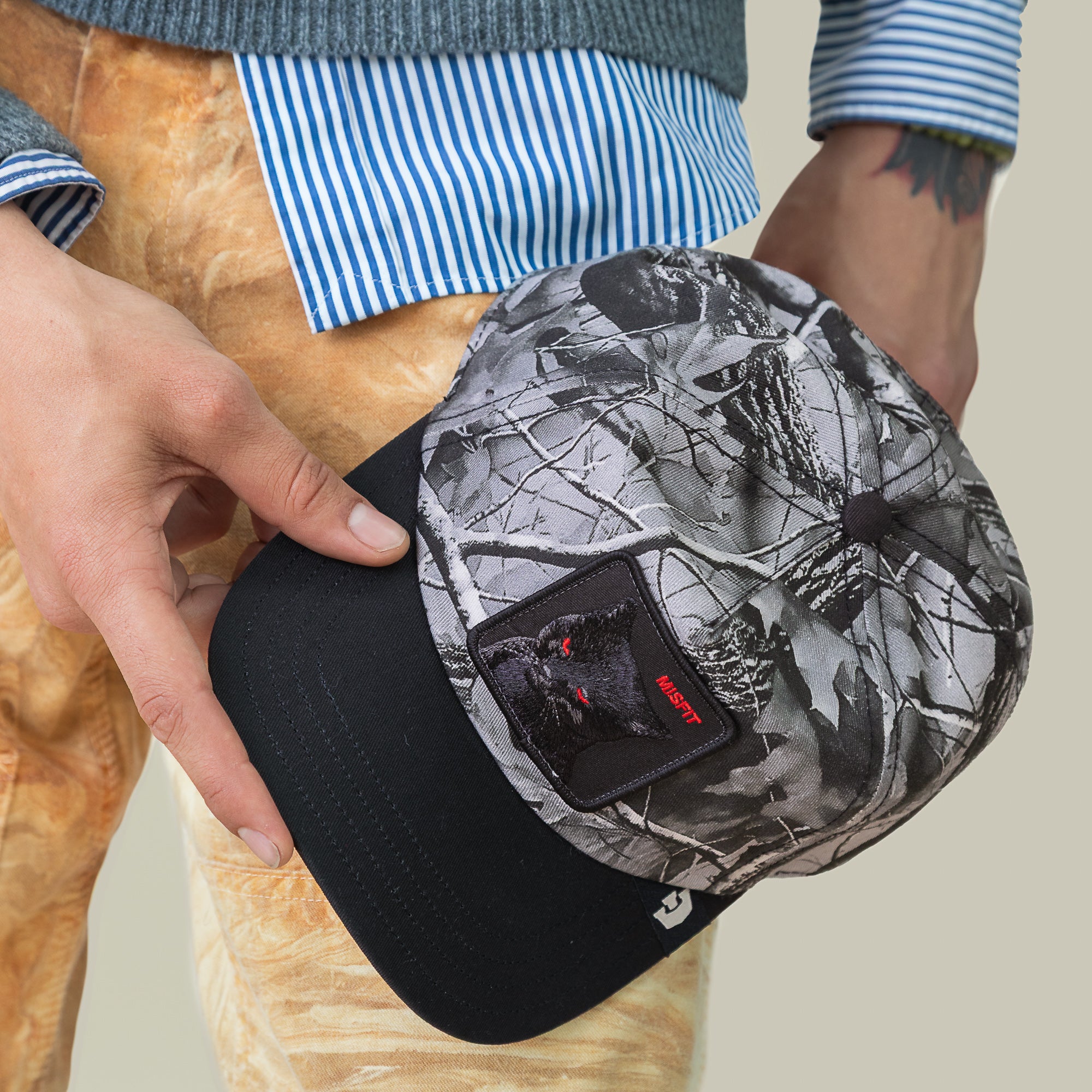 Someone holds a Goorin Bros. Misfit Camo baseball cap with an OBEY patch, dressed in a striped shirt, gray sweater, and patterned pants.