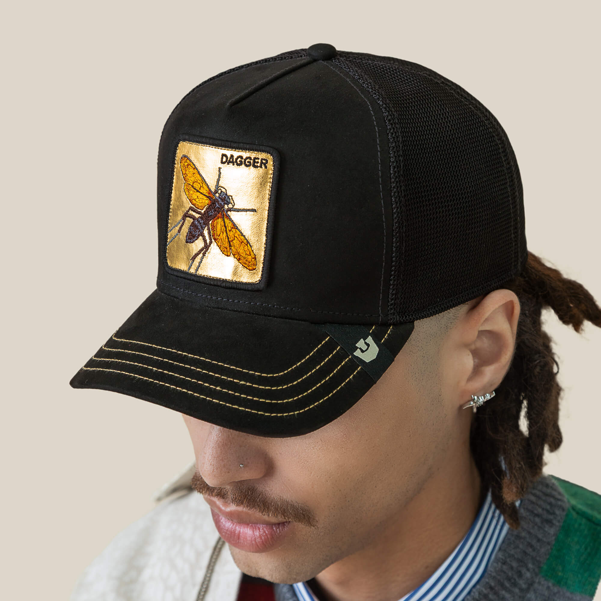 A person with earrings and a mustache wears the Goorin Bros. Golden Dagger black mesh trucker hat, featuring a sharp yellow insect patch on the front.