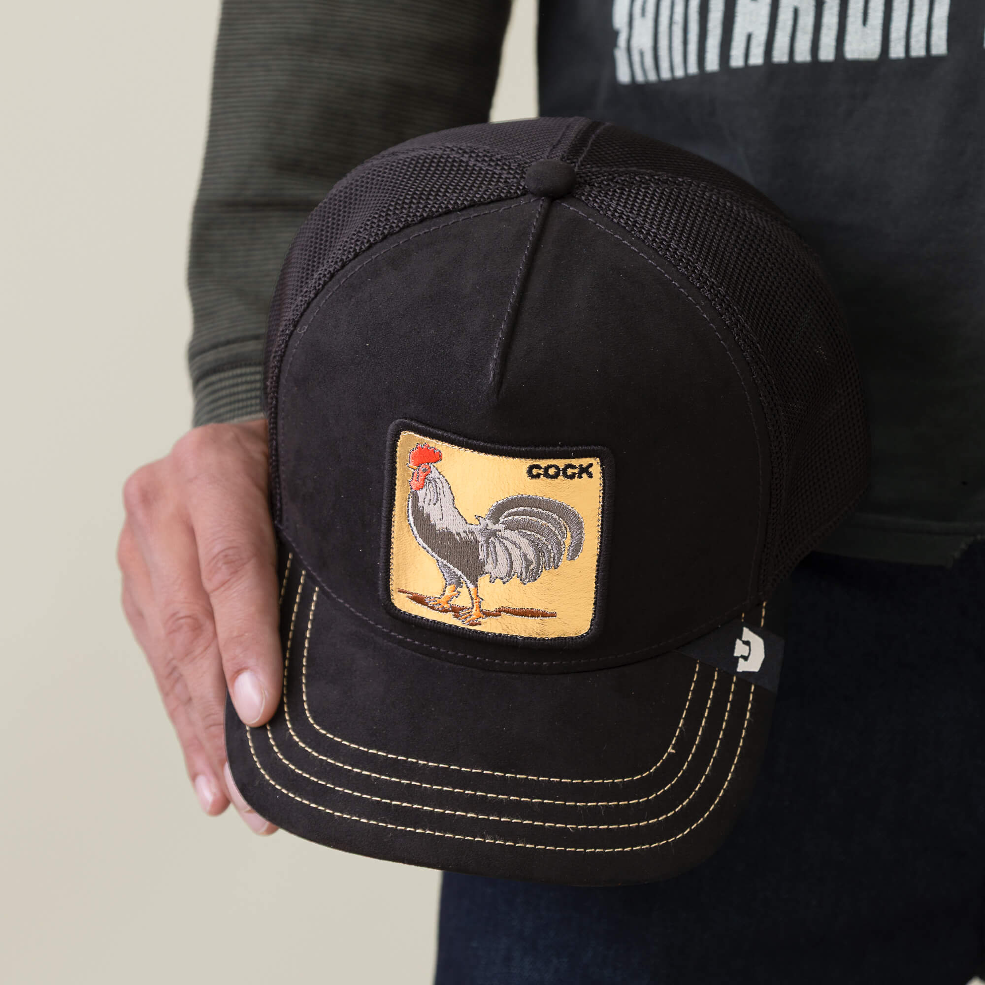 A person holding a black Goorin Bros. trucker hat called "Golden Cock," featuring a golden rooster patch with the word COCK above it.