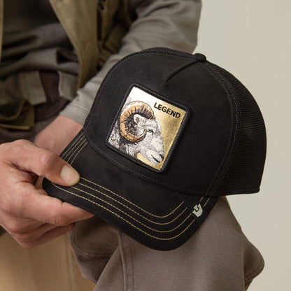 A person holds the Goorin Bros. Golden Legend cap—a black mesh baseball cap with gold stitching and a patch featuring a ram's head and "Golden Legend" above it, perfect for those with fearless style.