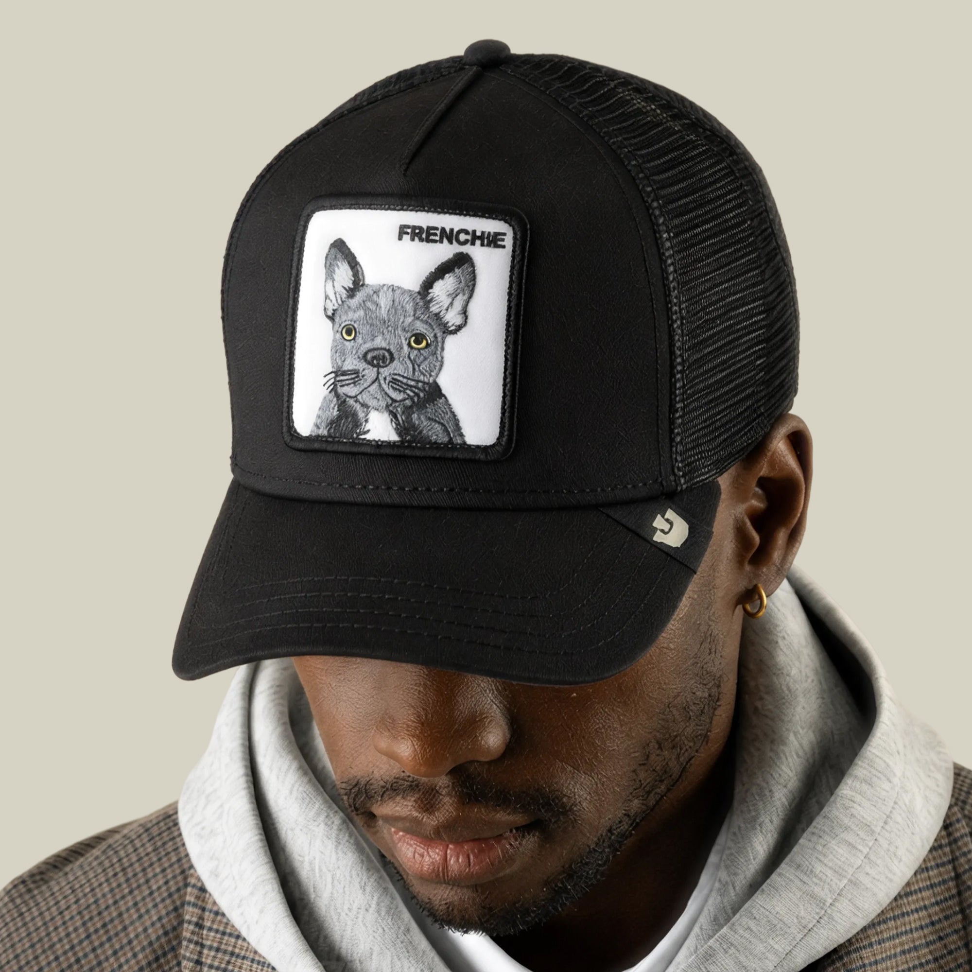 A person wearing a cute hat—a black Goorin Bros. Frenchie Trucker cap with a patch featuring an illustrated Frenchie and the word “FRENCHE”—dressed in a gray hoodie and brown plaid jacket.