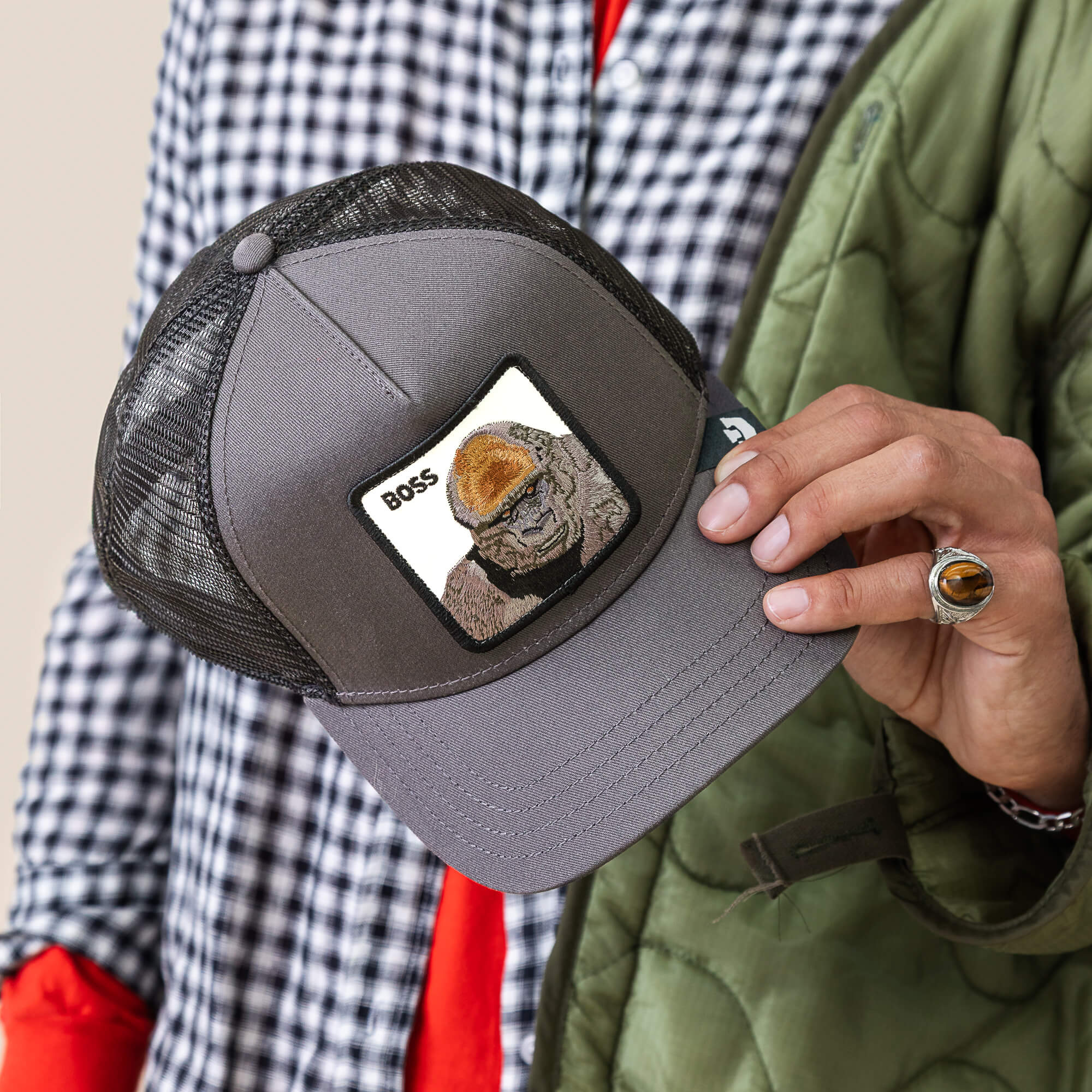 A person holds the Goorin Bros. Boss Trucker, a gray mesh hat with a bird BOSS patch, while wearing a checkered shirt, green jacket, and a brown stone ring.