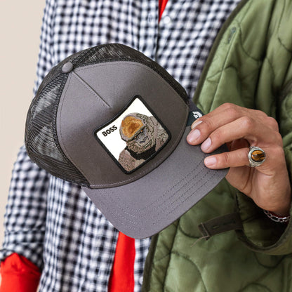 A person holds the Goorin Bros. Boss Trucker, a gray mesh hat with a bird BOSS patch, while wearing a checkered shirt, green jacket, and a brown stone ring.