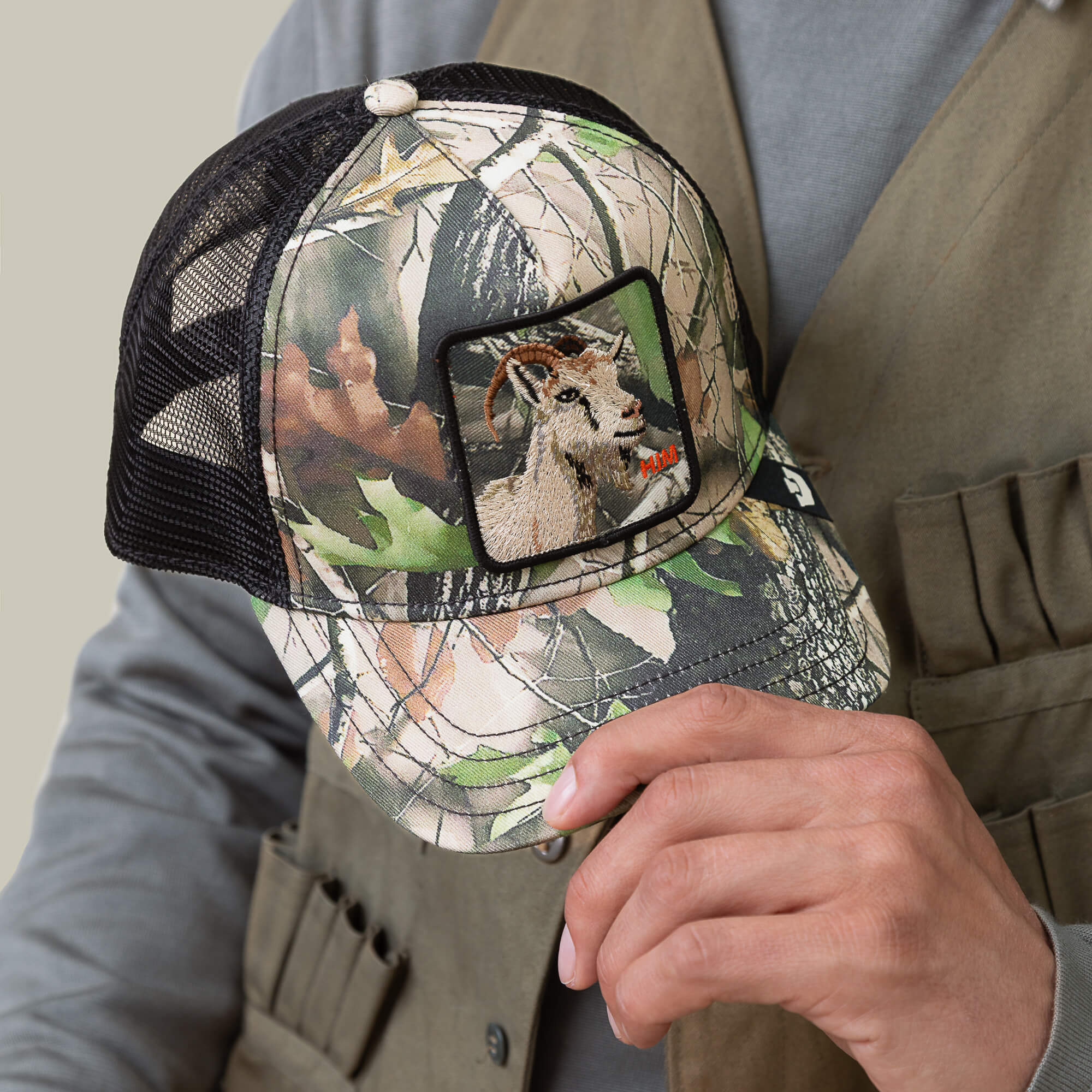 A person holds the Goorin Bros. Camouflage Goat hat, forest edition, featuring an embroidered goat patch on the front, while wearing a khaki vest.